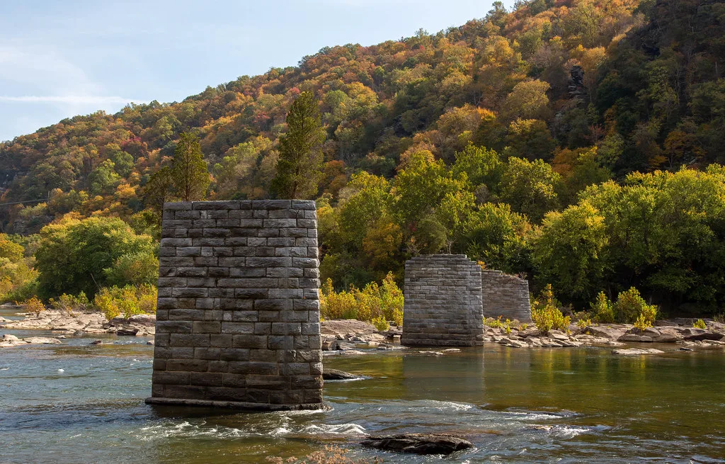 Trois piles de pierre d’un ancien pont sans tablier dans une rivière bordée d’une colline aux couleurs automnales.