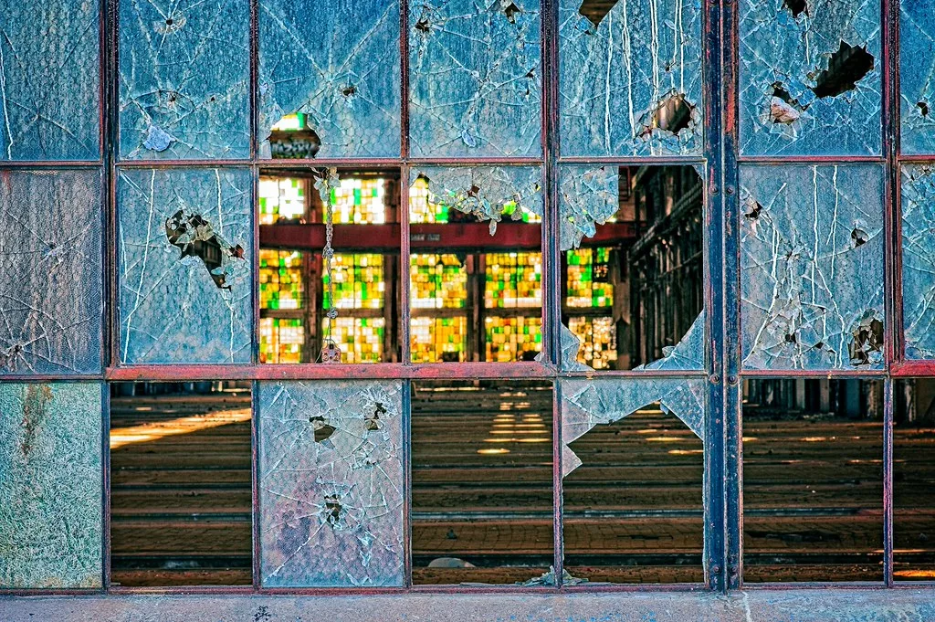 Shattered blue-tinted glazing in front of an abandoned room with colored stained glass.