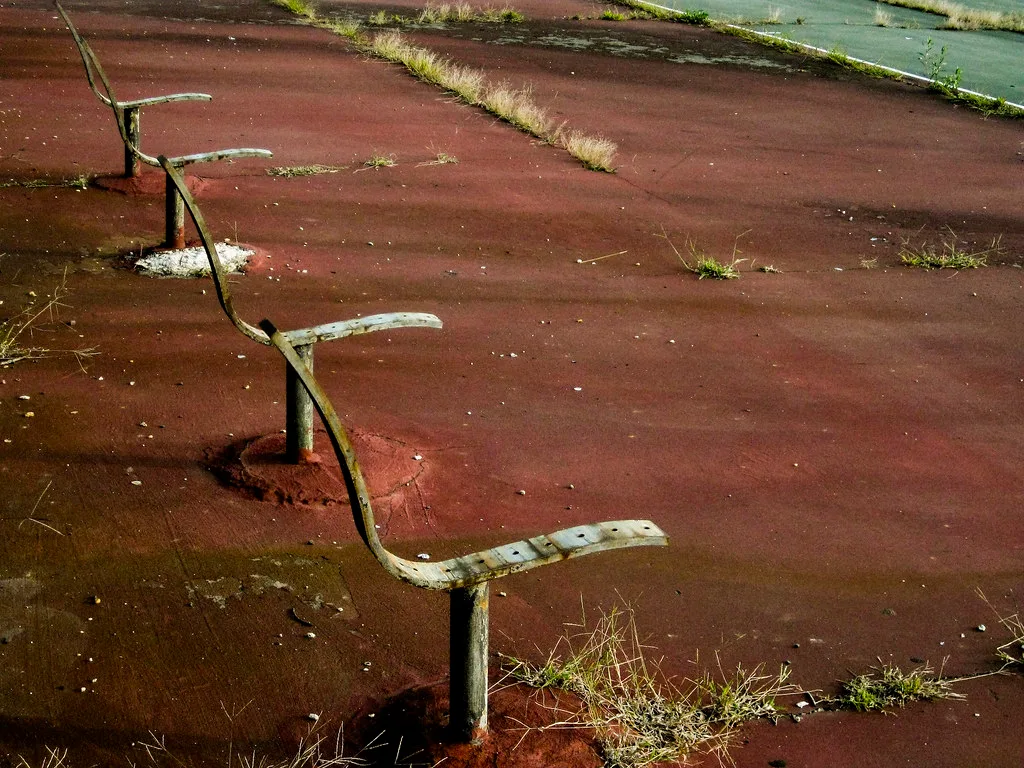 Row of rusty metal supports without a bench on a cracked, weedy red surface.