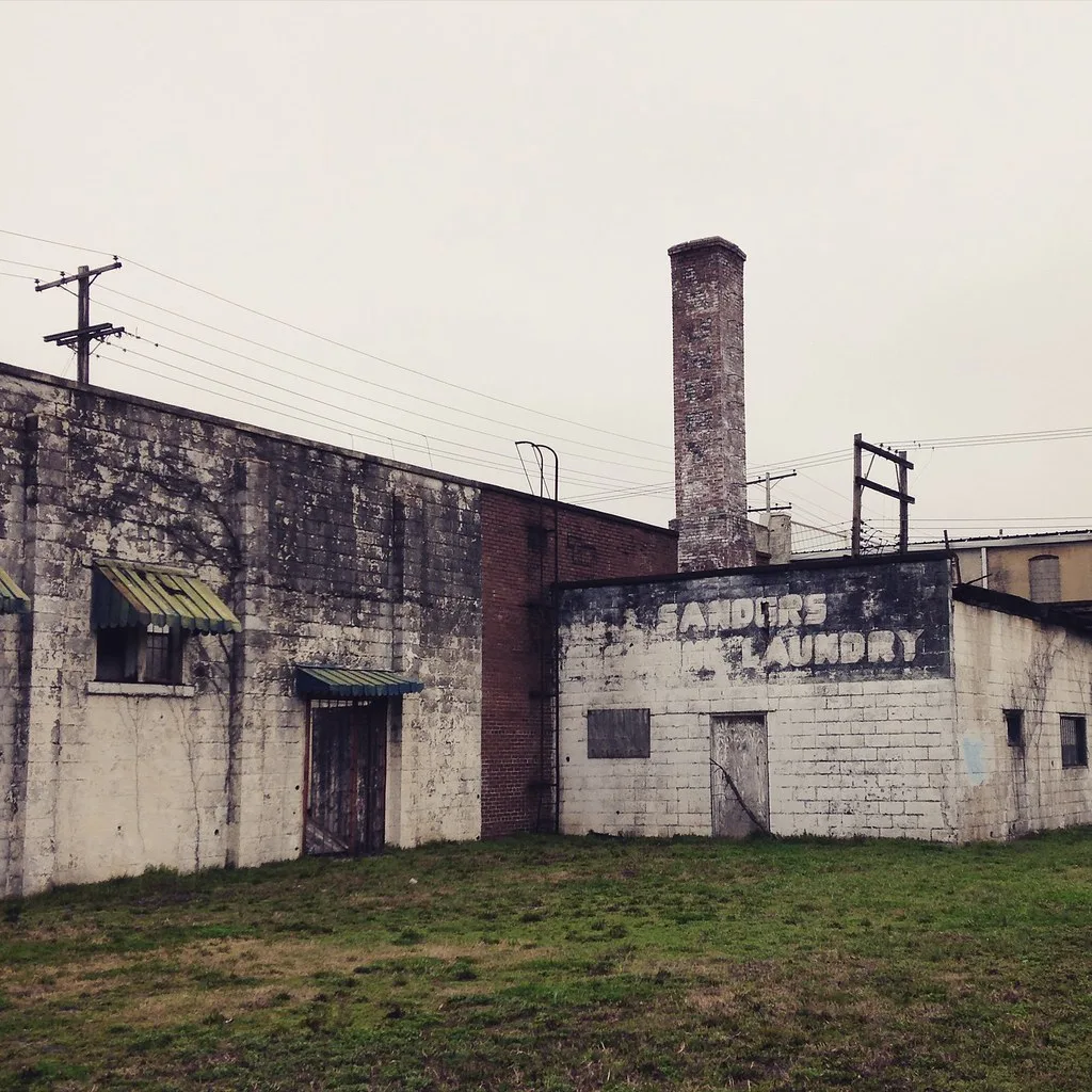 Abandoned former laundry with wood-covered doors and faded signage beneath a tall brick chimney.