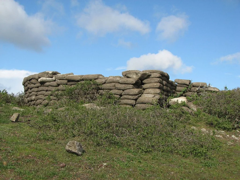 Ancien parapet en blocs de béton en forme de sacs empilés, partiellement envahi de broussailles sur une butte.