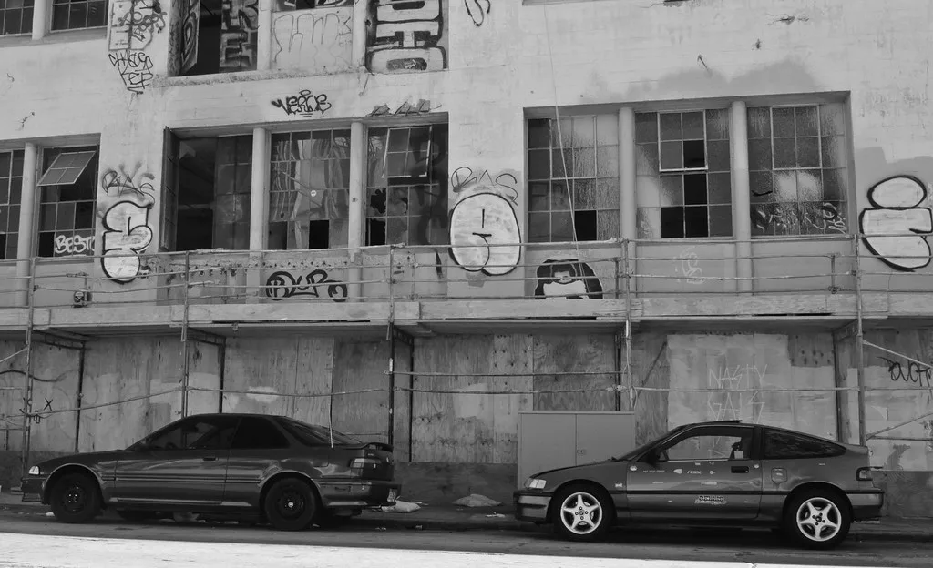 Façade abandonnée graffitée aux grandes fenêtres brisées et rez-de-chaussée condamné, derrière deux voitures en noir et blanc.