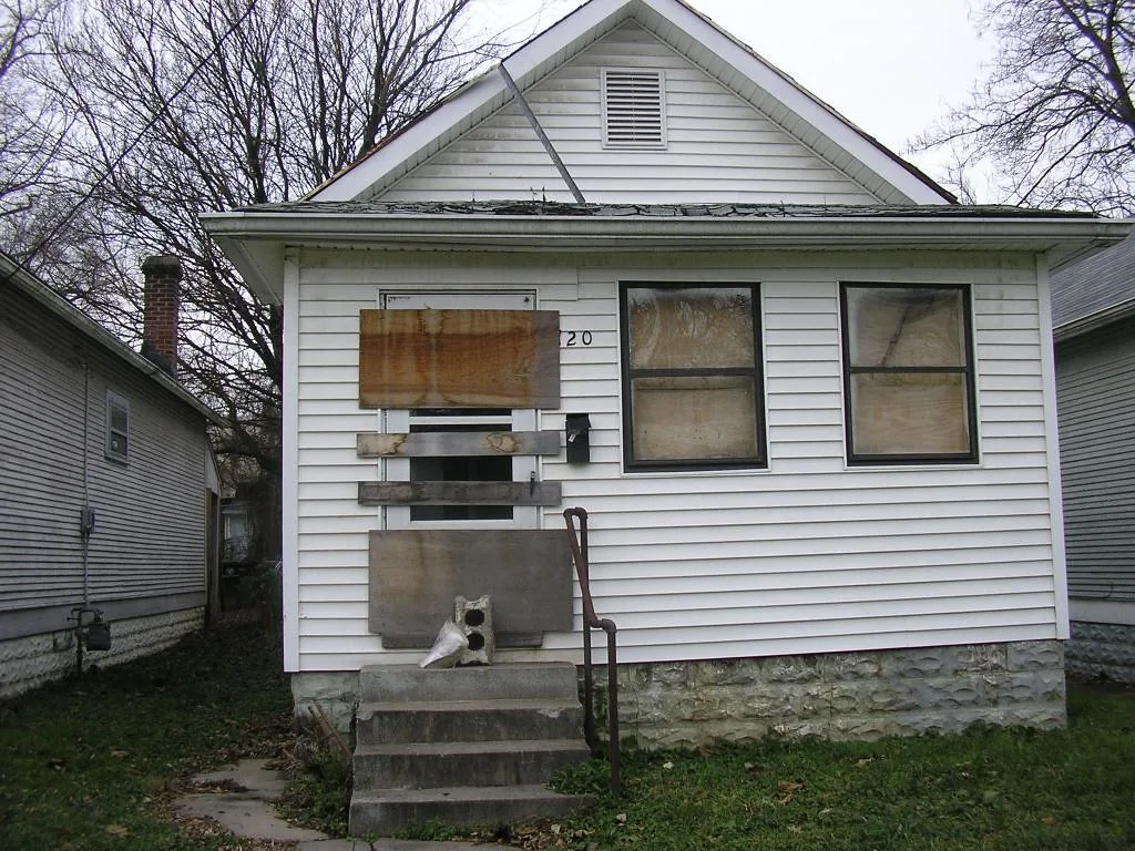 Small abandoned white house with covered windows, a plank-barred door and a deteriorated porch roof.