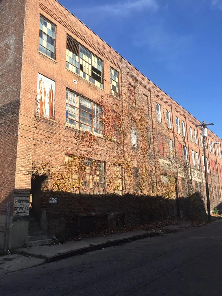 Longue façade d’usine en brique avec fenêtres condamnées et lianes desséchées.