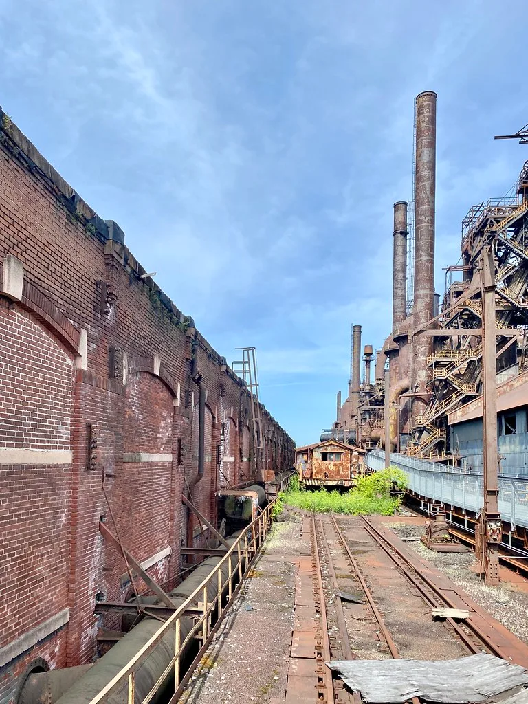 Voies rouillées et ancien wagon entre un mur de brique et de hautes cheminées industrielles.