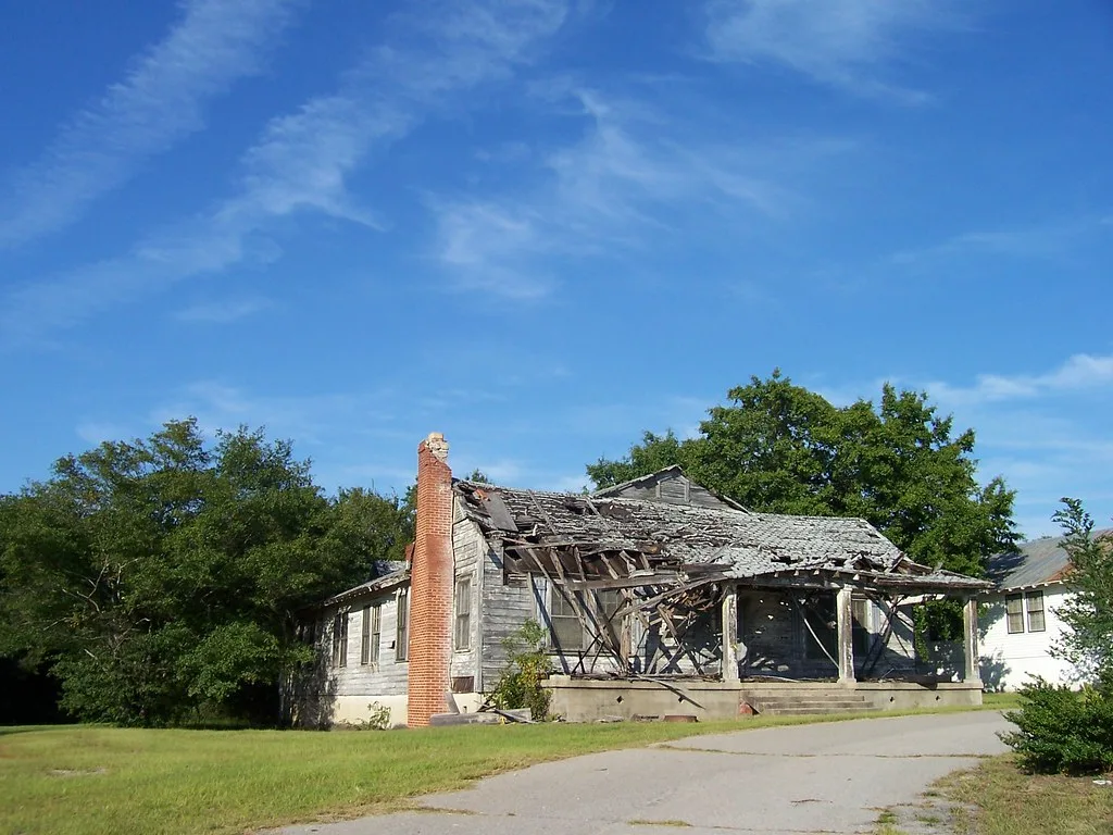 Maison abandonnée de bois gris au toit et porche effondrés, avec haute cheminée de briques au bord d’une allée.