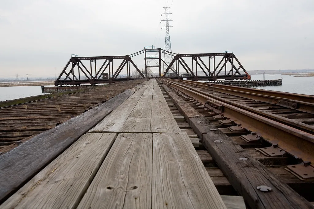 Abandoned railway bridge with deteriorated wooden decking and a rotated steel span interrupting the rails above the water.