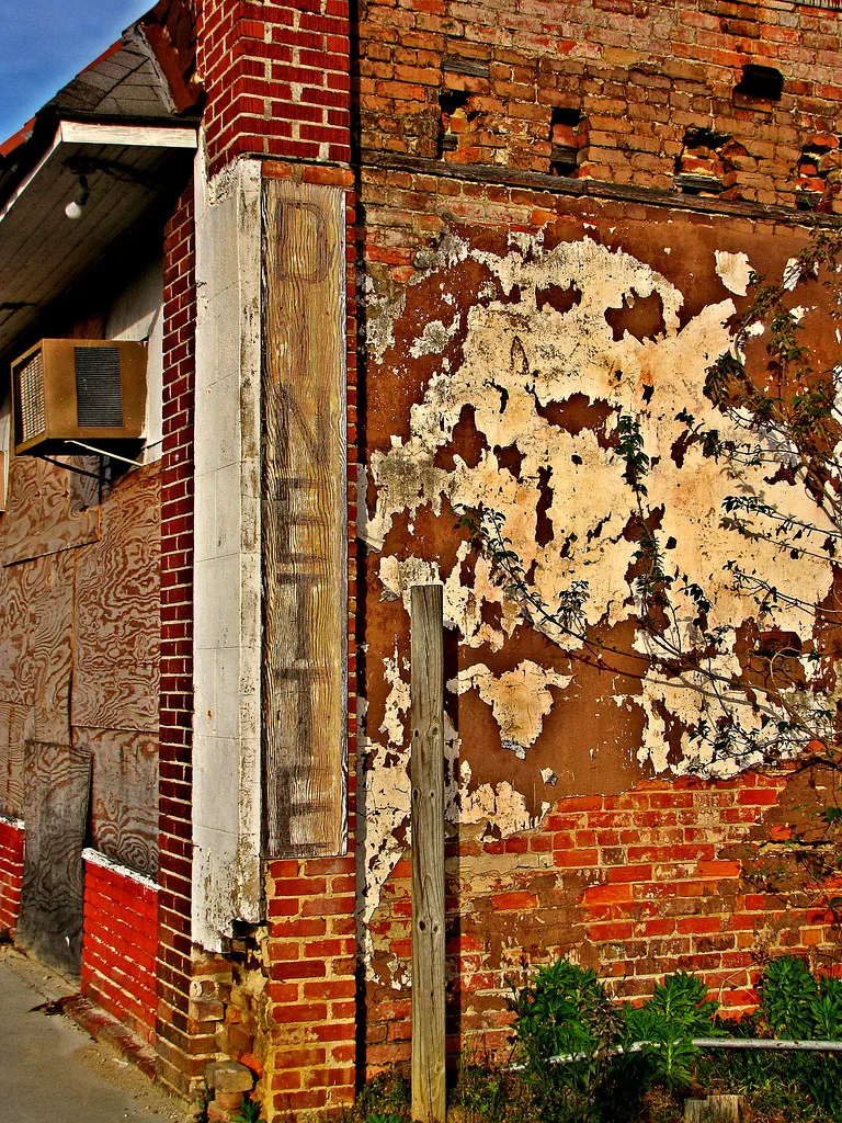 Angle d’un bâtiment de briques abandonné aux ouvertures couvertes de bois et à l’enduit écaillé envahi de plantes.