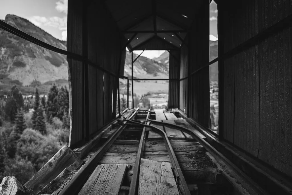 Anciens rails dans une galerie de bois et métal ouverte sur les montagnes, en noir et blanc.