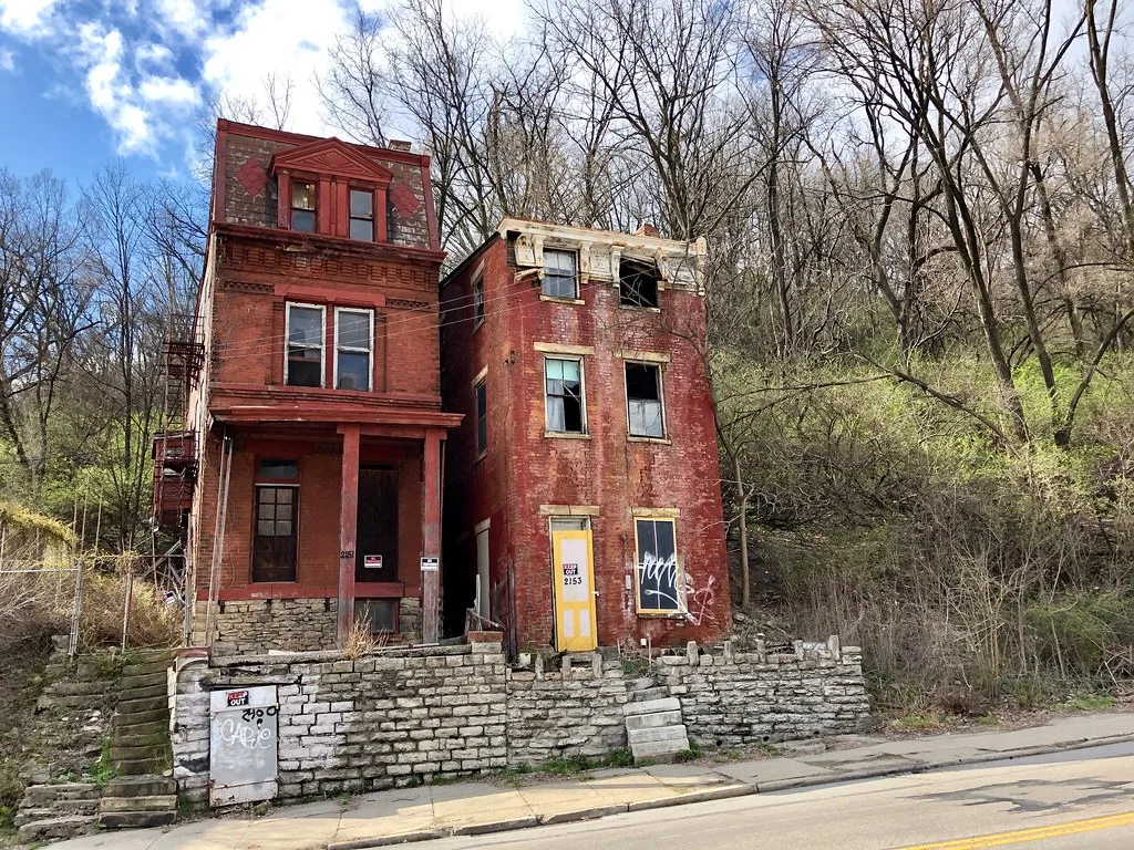 Two old brick houses with broken windows at the foot of a wooded slope.