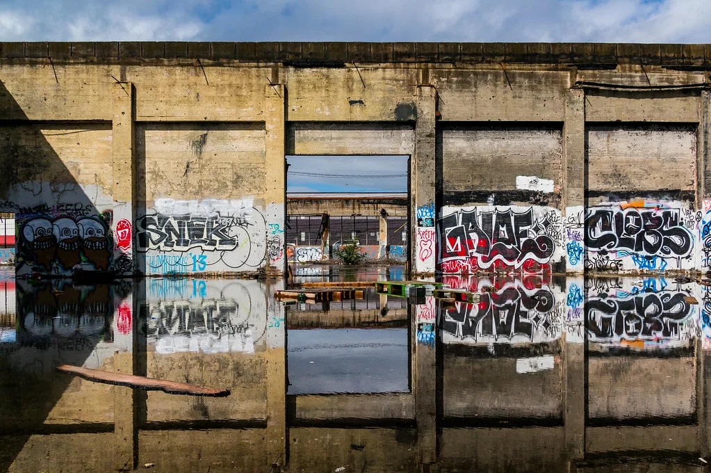 Murs de béton tagués d’une ruine industrielle abandonnée se reflétant dans l’eau au sol sous le ciel ouvert.