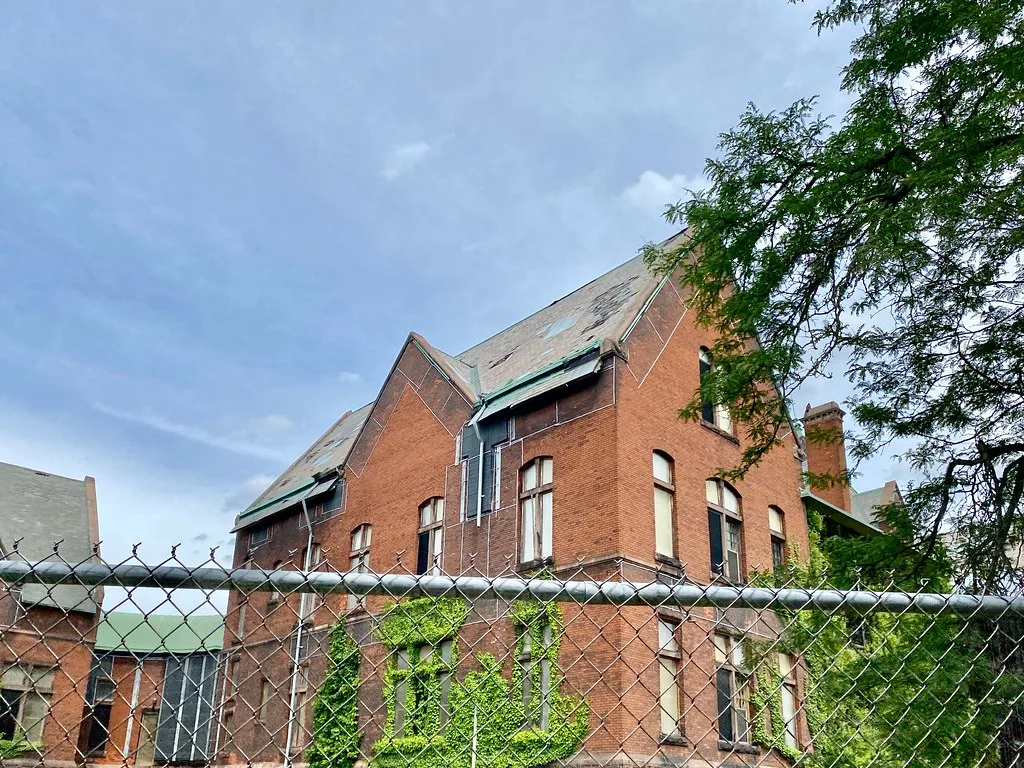 Old brick building with boarded openings behind a wire fence.