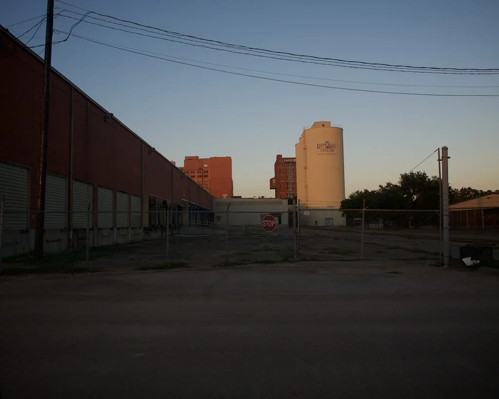 Cour délaissée d’une usine avec grand silo clair, entrepôts fermés et végétation dans le bitume.