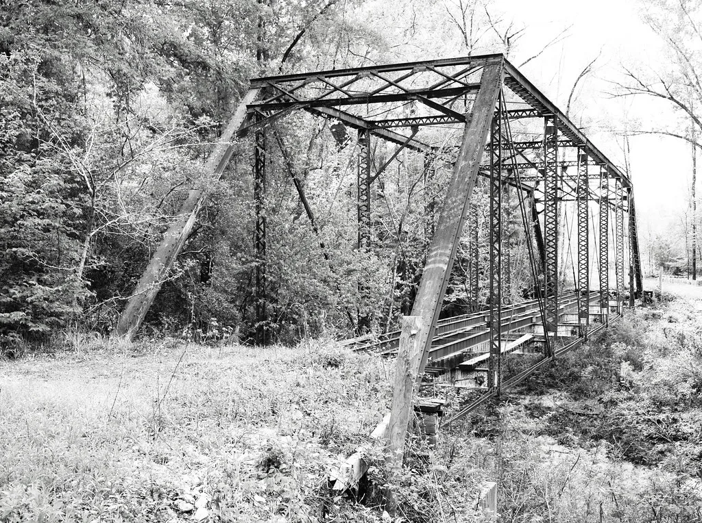 Old metal bridge without decking among trees.