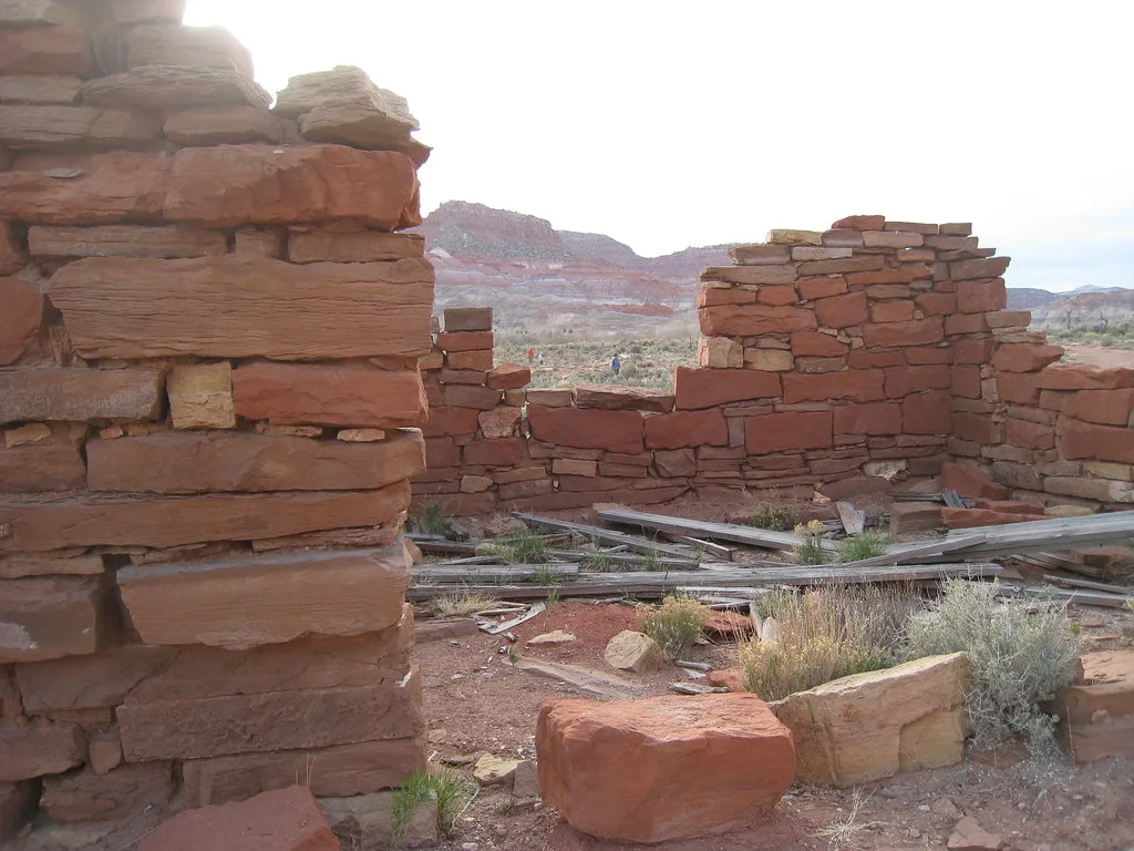 Ruines de murs en grès rouge et planches au sol dans un paysage aride.