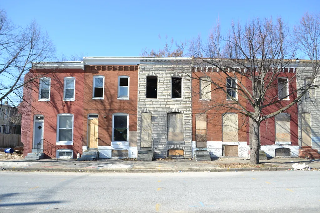 Maisons mitoyennes abandonnées de brique et pierre claire aux fenêtres vides ou condamnées le long d’une rue.