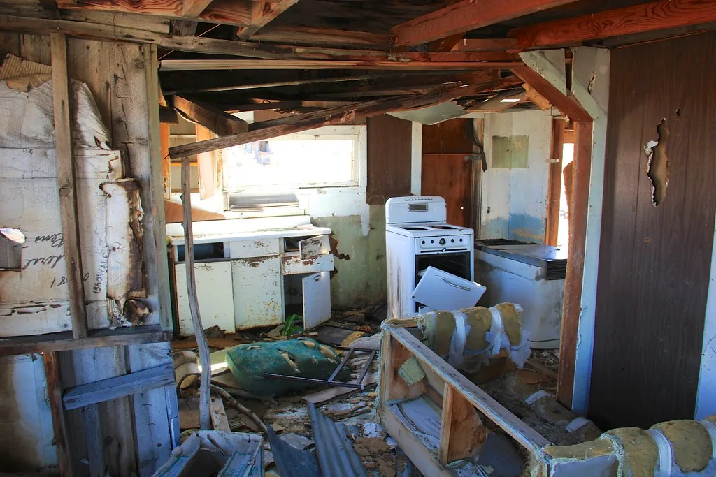 Abandoned kitchen with a white stove, broken cabinets and fallen beams amid debris.