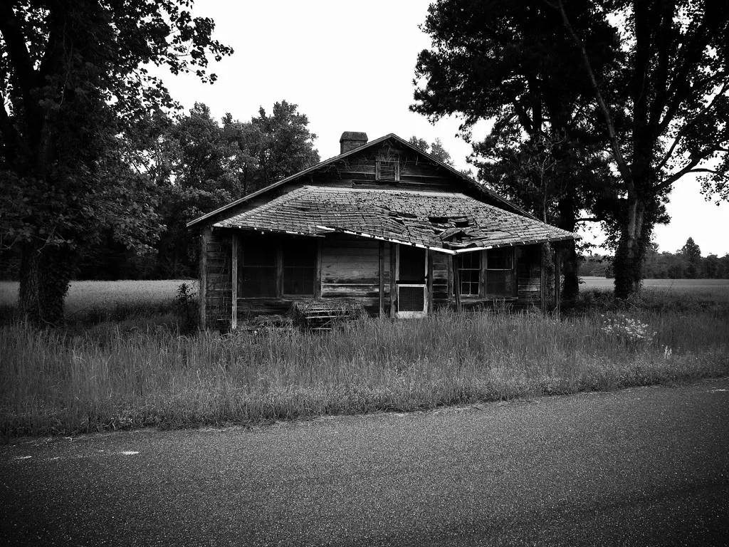 Old wooden house with a collapsed porch roof behind tall grass.