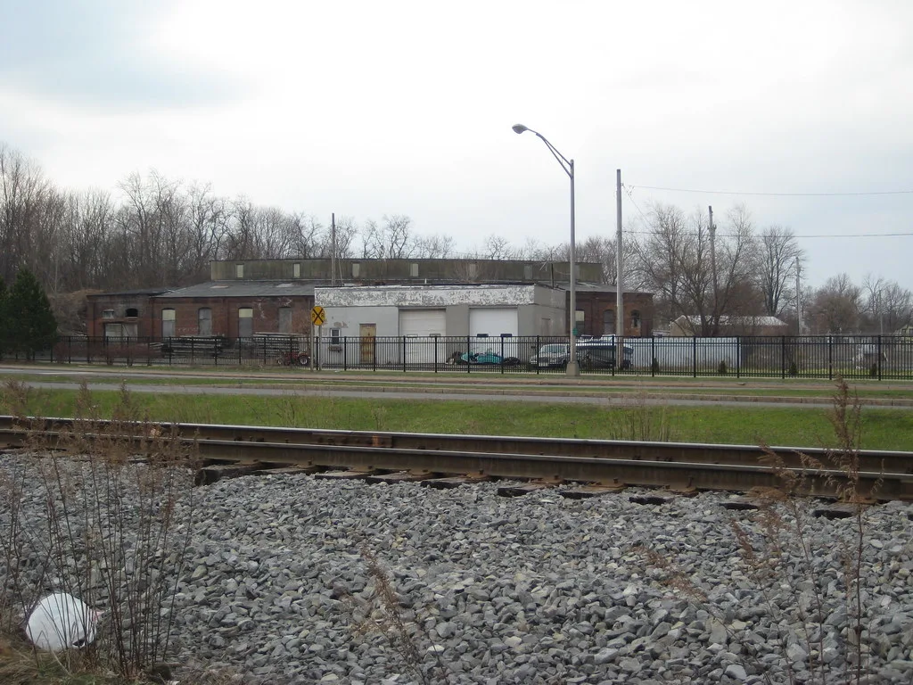 Industrial building with boarded openings behind a fence and railway track.