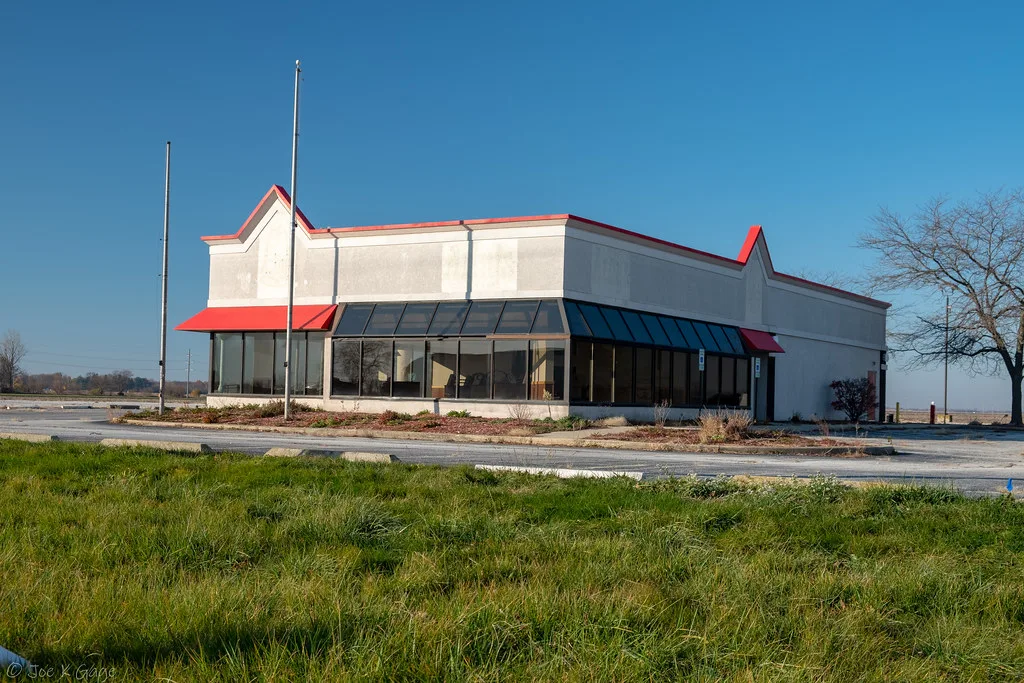 Ancien restaurant blanc et rouge vide devant un parking gagné par les herbes.