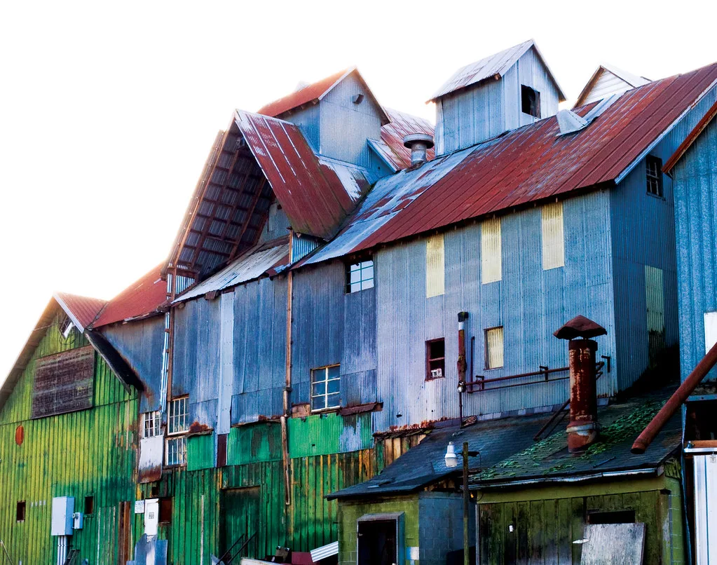 Old blue and green industrial building with red roofs and boarded openings.