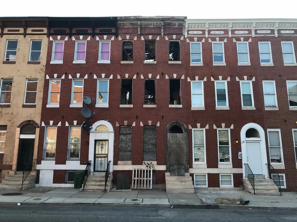 Abandoned brick townhouse with gaping windows and a boarded entrance between occupied facades.