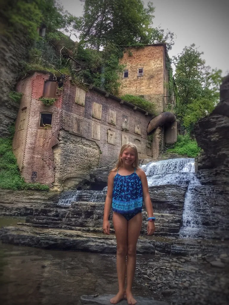 Abandoned old brick building with boarded windows and a rusty pipe beside a waterfall, with a child in the foreground.