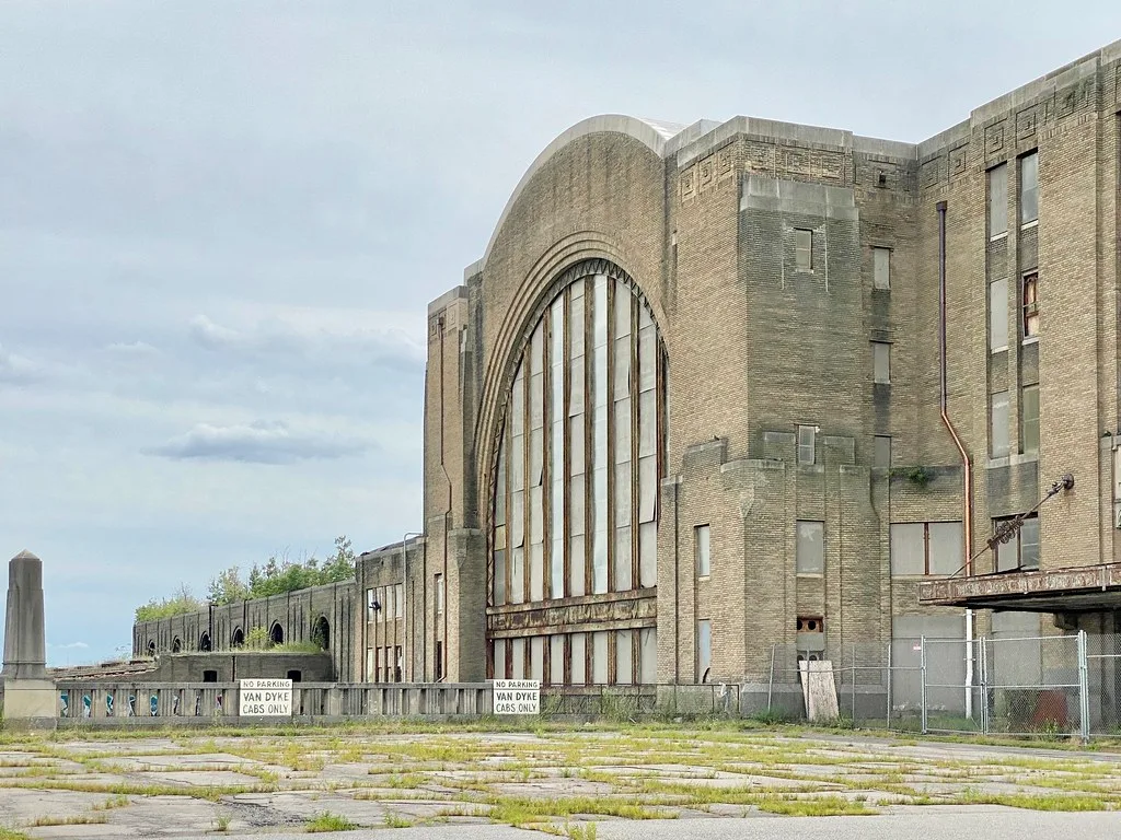 Ancienne gare aux fenêtres condamnées derrière un parvis envahi d’herbes.