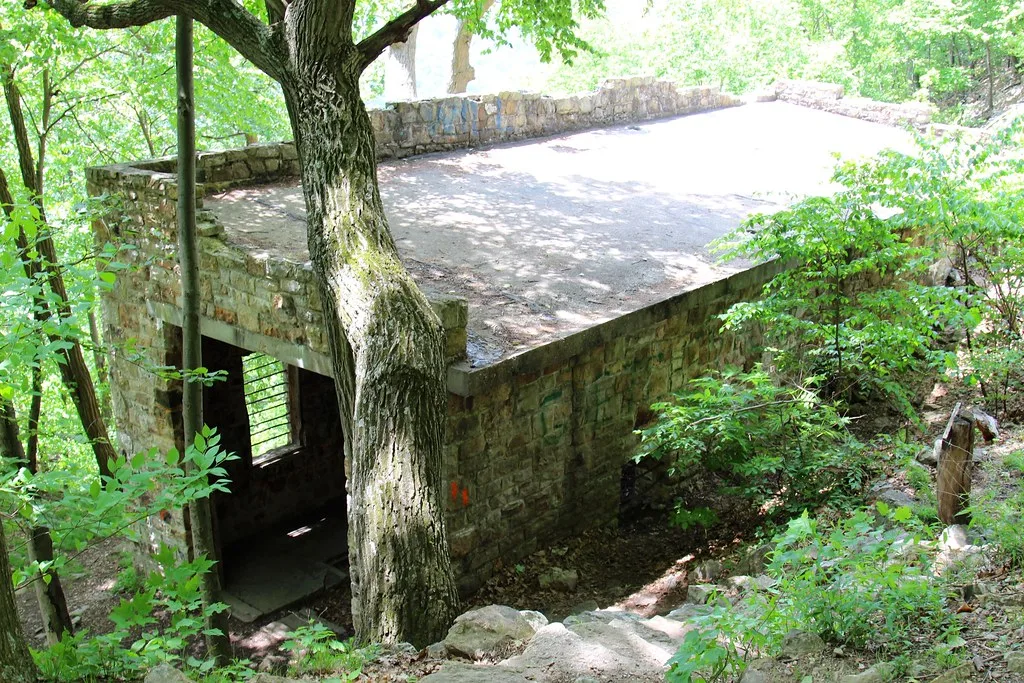 Petit bâtiment abandonné de pierre à toit plat et ouvertures vides, entouré d’arbres sur une pente rocheuse.