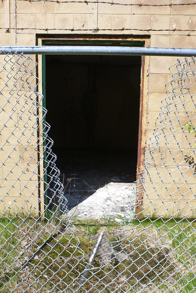 Dark opening in a beige wall behind a cut fence, with rubble on the threshold.