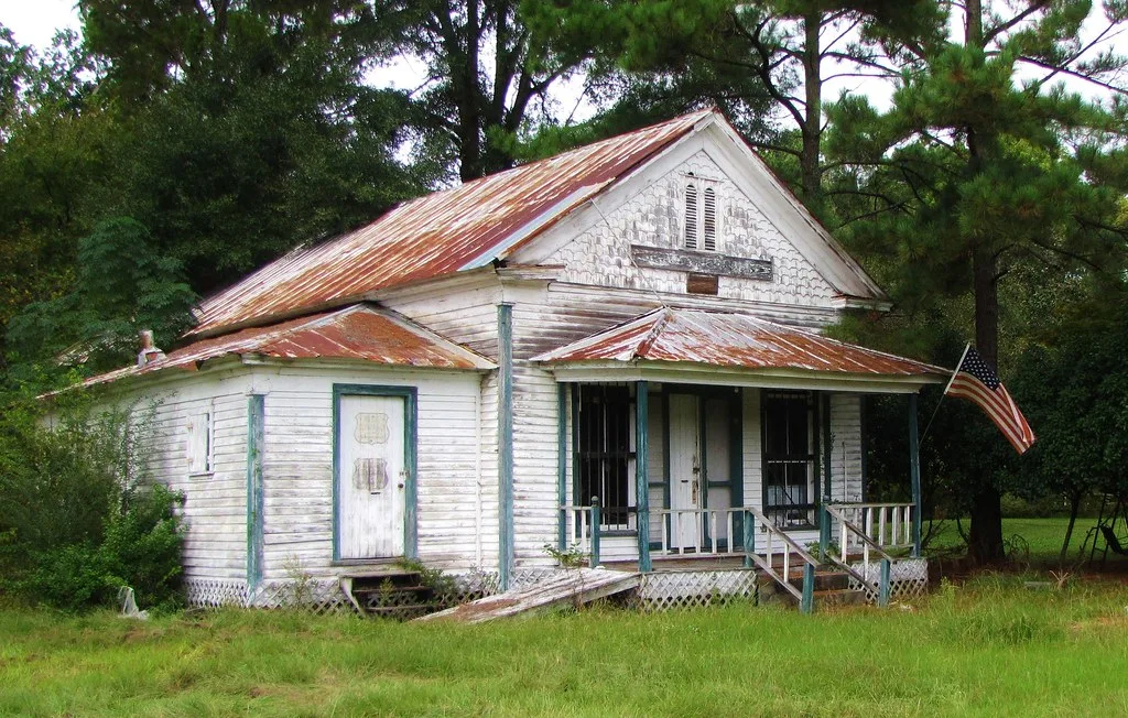 Petite maison blanche délaissée à toiture rouillée, porche dégradé et portes condamnées dans les hautes herbes.