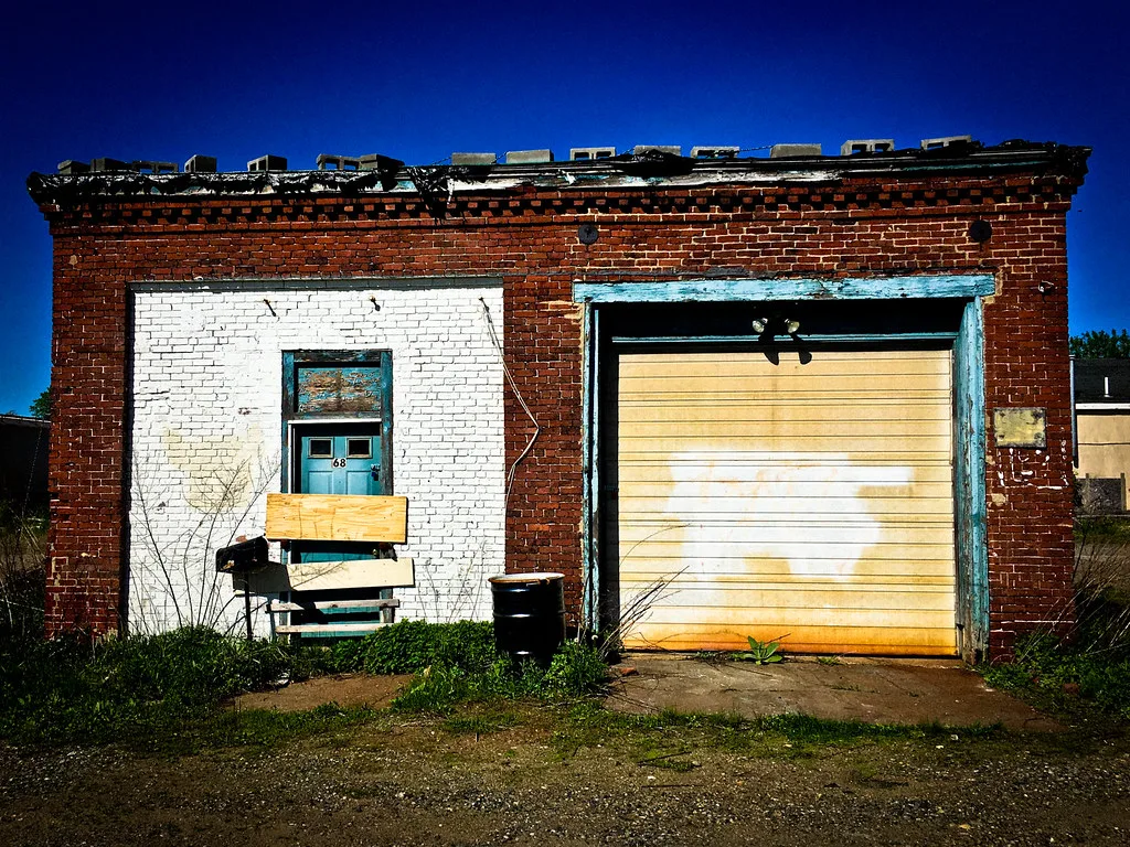 Neglected small brick garage with a plank-barred side entrance and a large yellow door facing weeds.