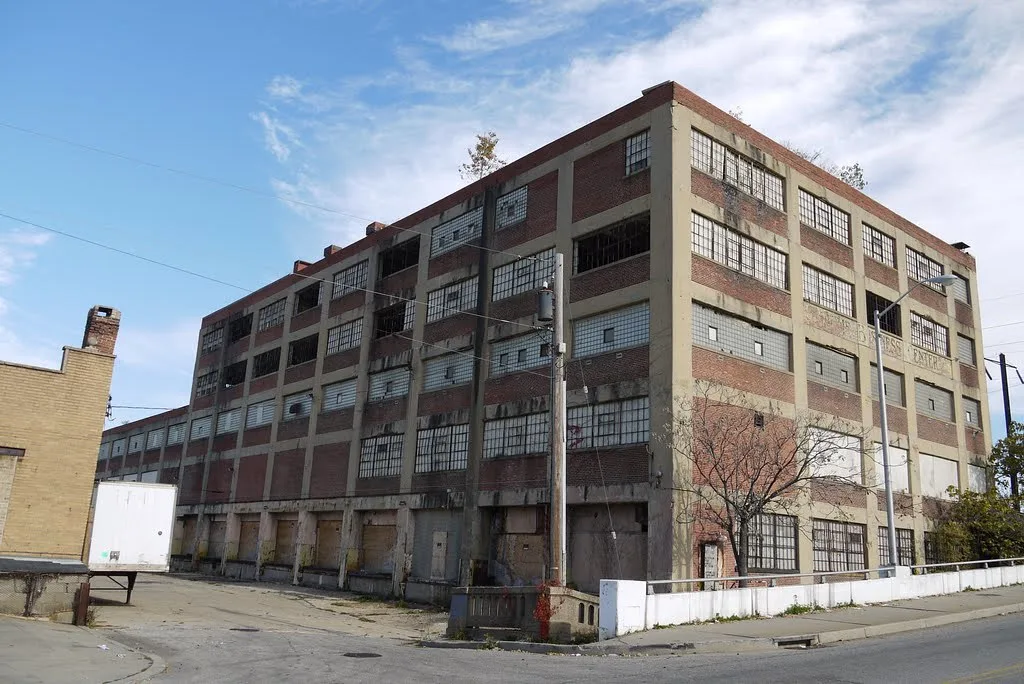 Large abandoned brick-and-concrete factory with broken windows, sealed loading bays and trees growing on the roof.