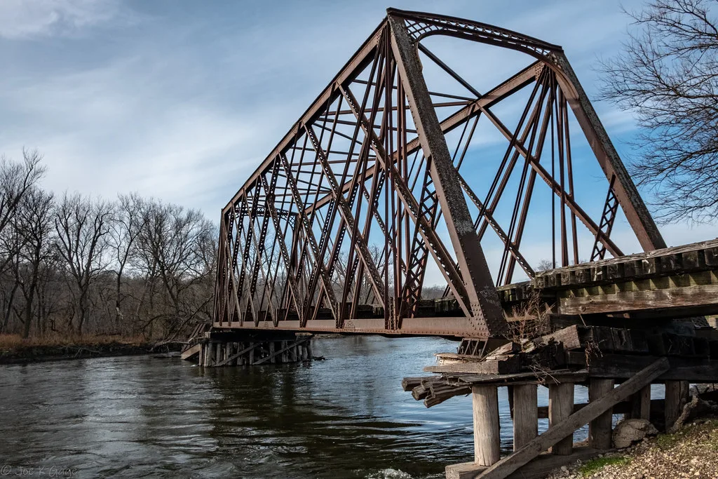Old metal railway bridge on decayed wooden supports above a river.