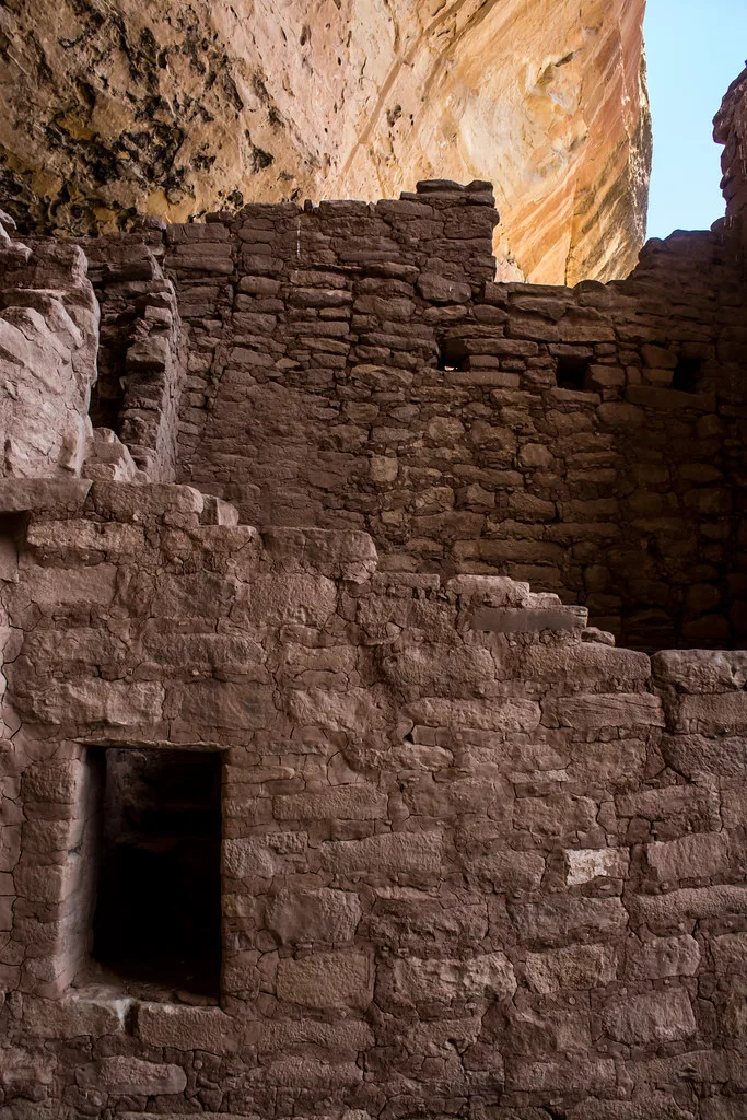 Ruins of stone dwellings with small rectangular openings beneath an ochre cliff.