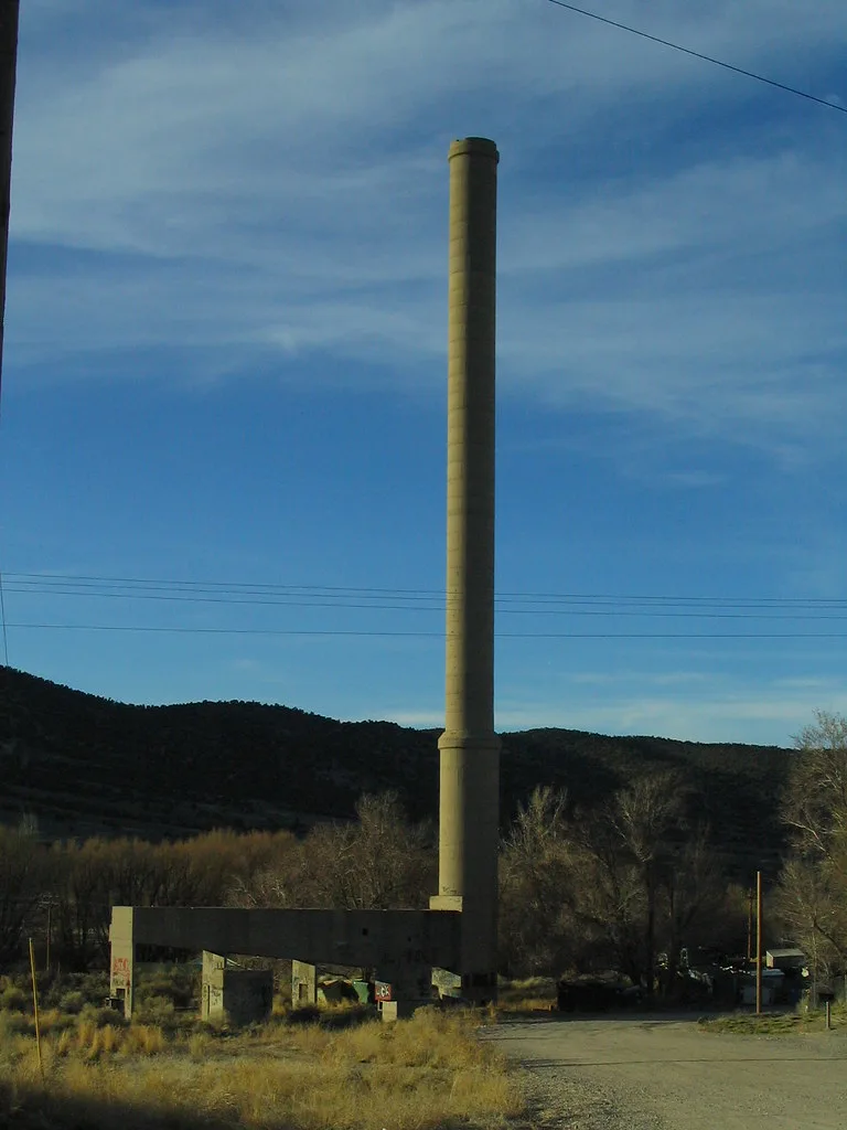 Tall abandoned industrial chimney above remaining concrete pillars and beams on grassy ground.
