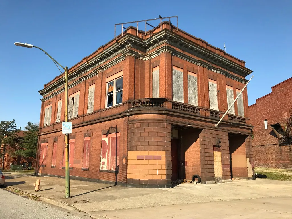 Two-storey abandoned red brick building with boarded or broken windows and a deteriorated cornice on a street corner.