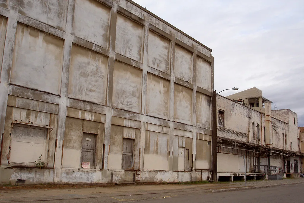 Longue façade d’usine abandonnée claire aux baies condamnées, auvent de quai dégradé et tour aux ouvertures vides.