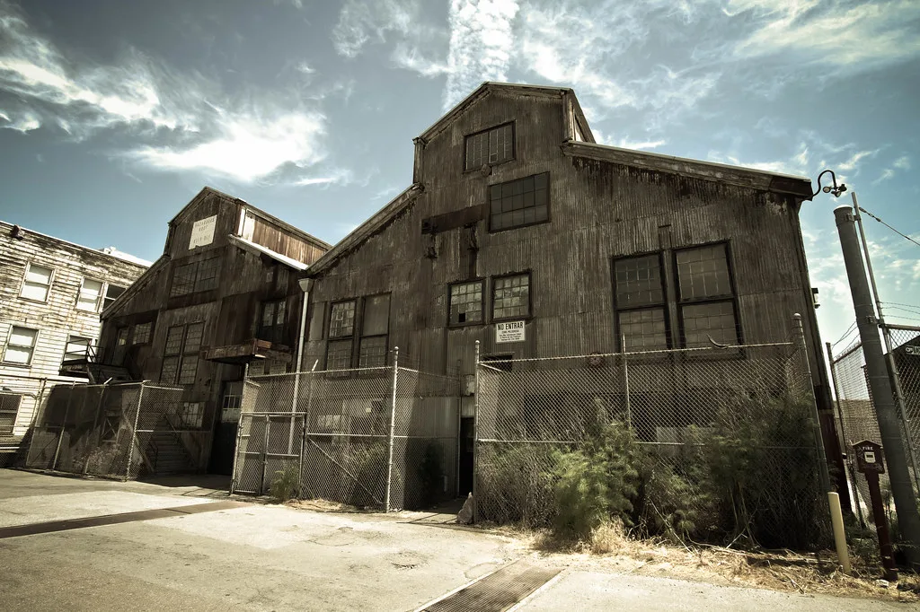 Abandoned former workshops with tall deteriorated metal-clad gables and weathered windows behind a fence.