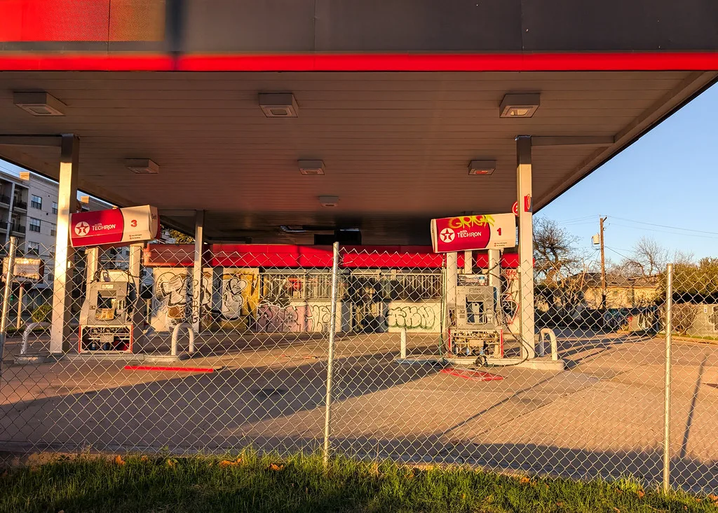 Abandoned gas station behind a fence with two graffitied pumps and a shop with broken windows.
