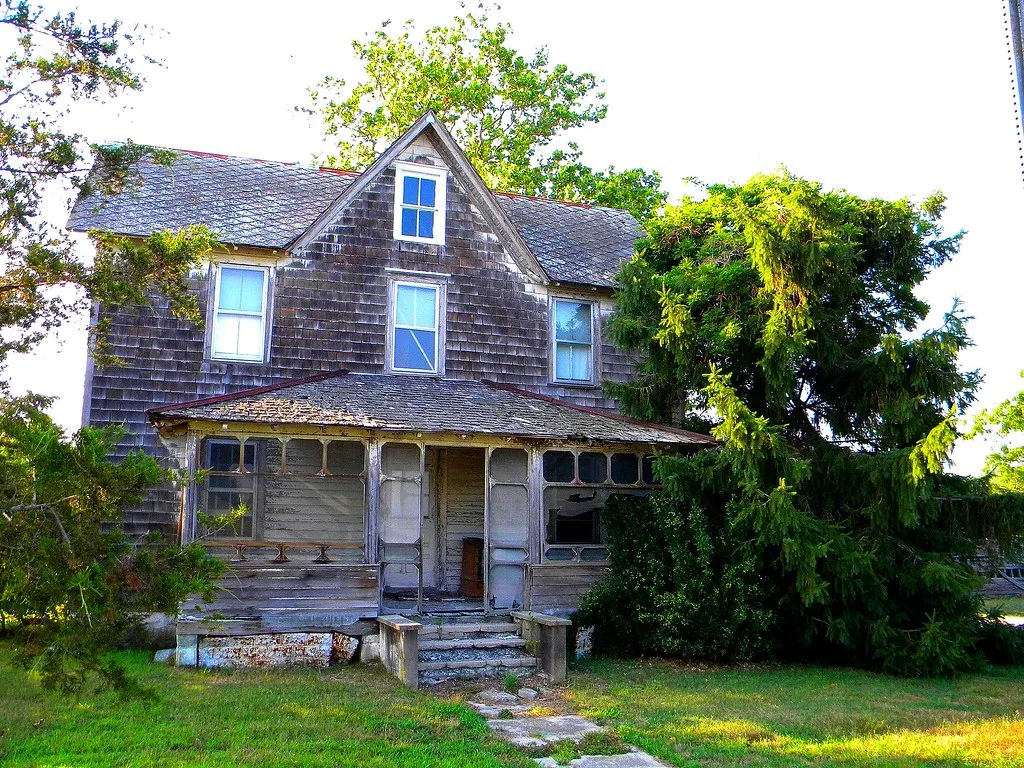 Old wooden house with a deteriorated porch partly covered in vegetation.
