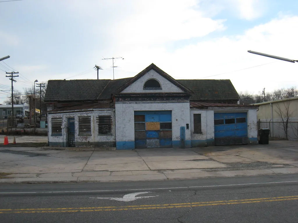 Ancien garage blanc et bleu aux fenêtres obturées et portes détériorées devant une aire fissurée.