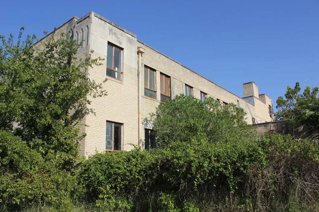 Ancien bâtiment en briques claires derrière des arbustes envahissants.