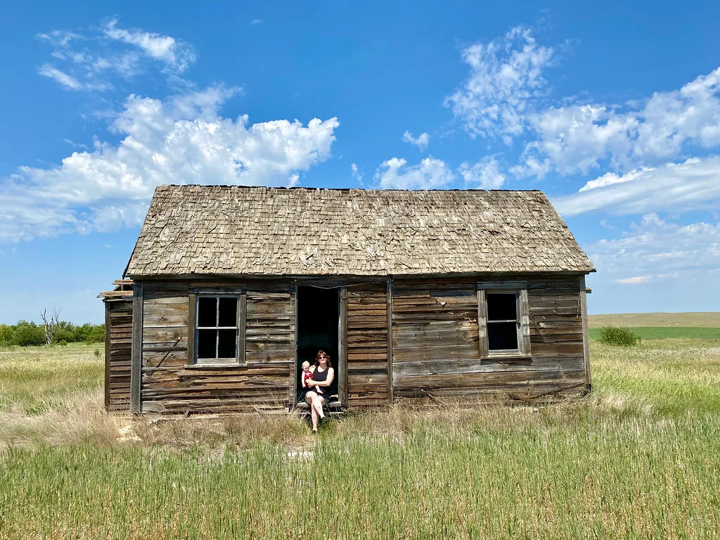 Abandoned wooden cabin with empty windows in a meadow, with an adult and child on the doorstep.