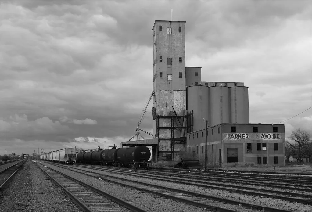 Old silo and deteriorated building beside railway tracks, in black and white.