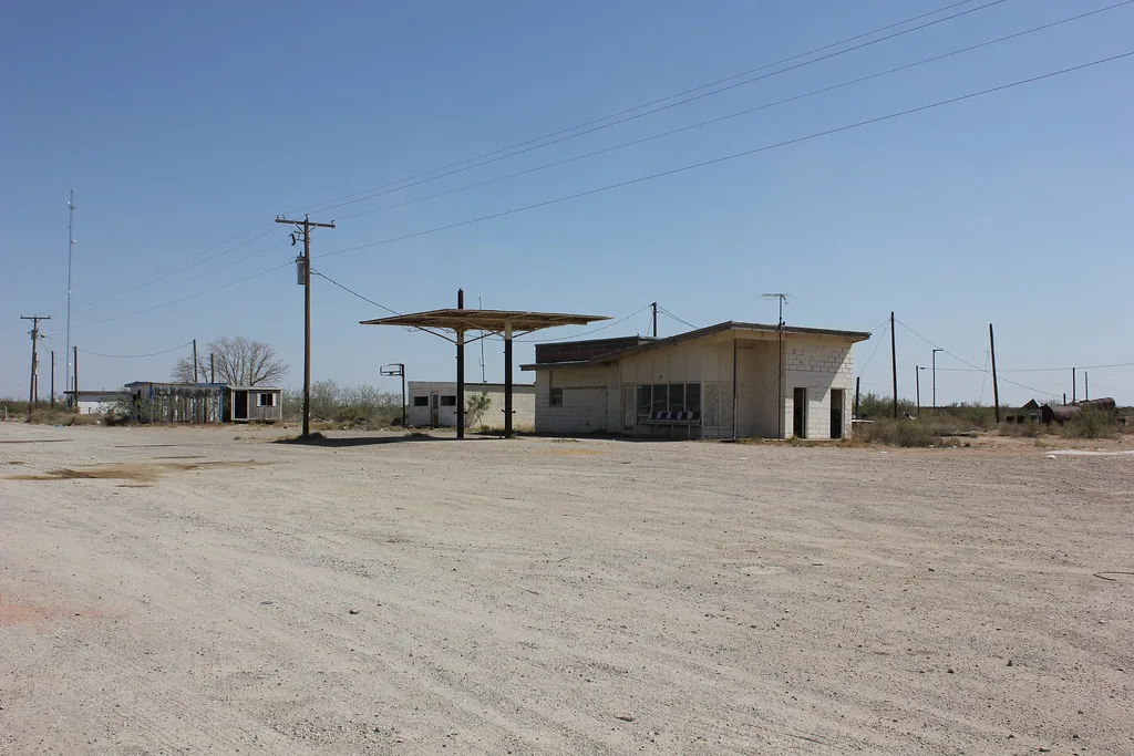 Station-service délaissée avec auvent vide et petit bâtiment clair au bord d’une grande aire déserte.