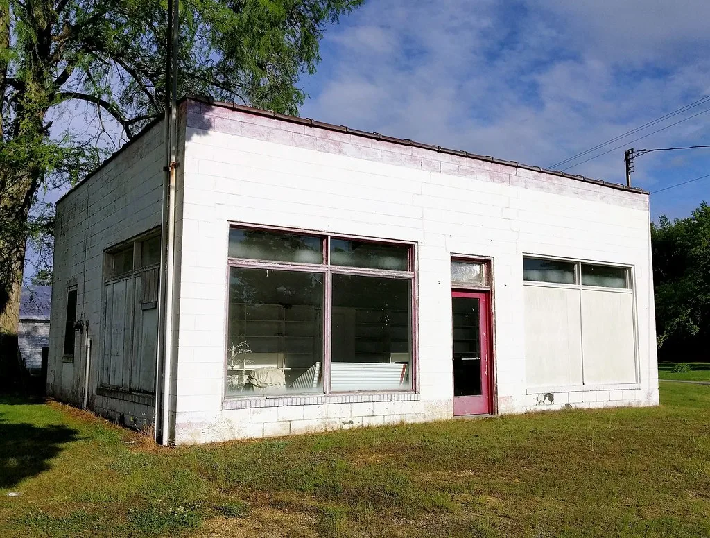 Small old white shop with partly boarded windows and a red door.