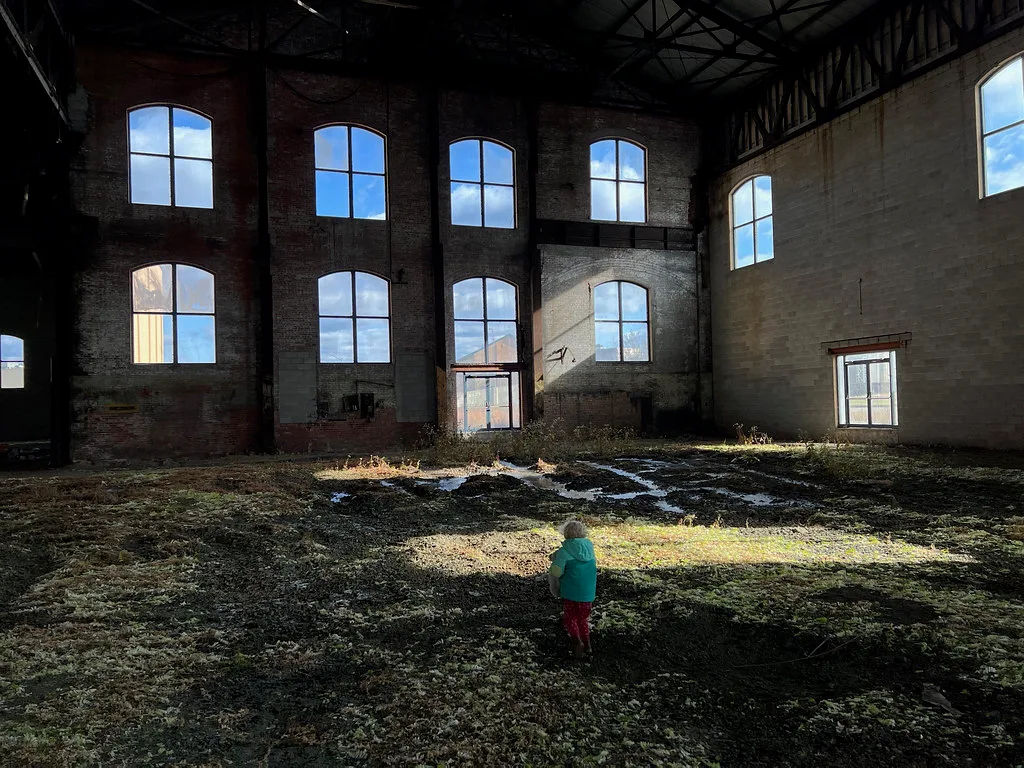 Grande halle abandonnée aux hautes fenêtres cintrées et murs de brique, avec mousses et plantes couvrant le sol irrégulier.