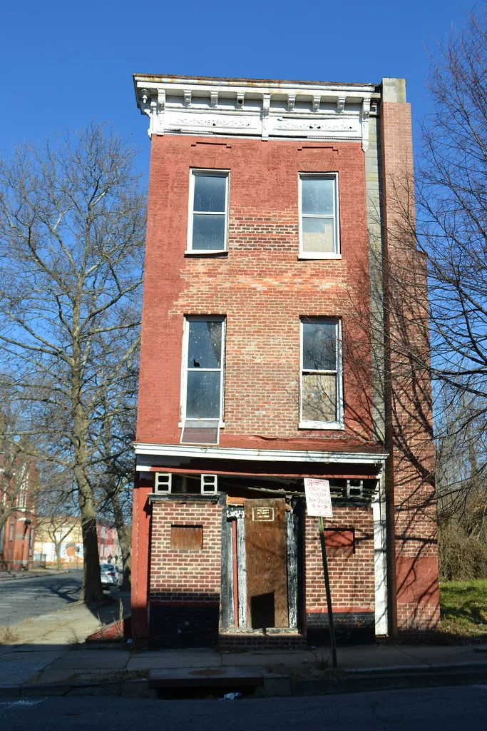 Narrow three-storey abandoned red brick building with broken windows and a boarded, deteriorated former shop entrance.