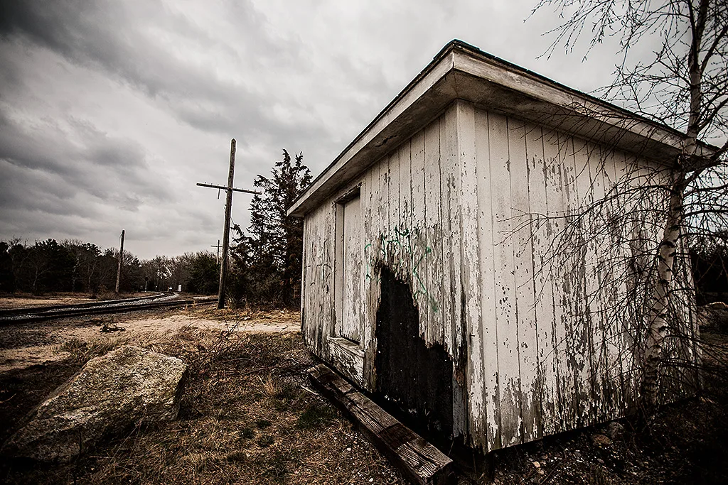 Small white shed with rotten, perforated cladding beside a railway track.